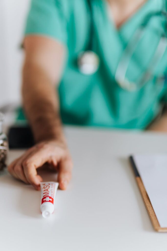 Unrecognizable man doctor wearing medical uniform offering medical ointment in tube while sitting at table with clipboard and stethoscope during work